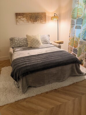 Ein Bett mit grauer Steudecke, ein weißer, flauschiger Teppich, eine Wandlampe und ein Gemälde über dem Bett.