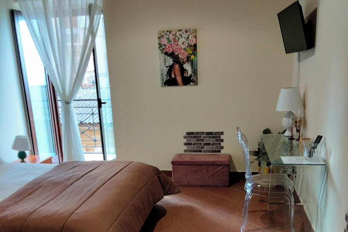 A bedroom with a bed, a desk, a chair, a TV, and a window with curtains.
