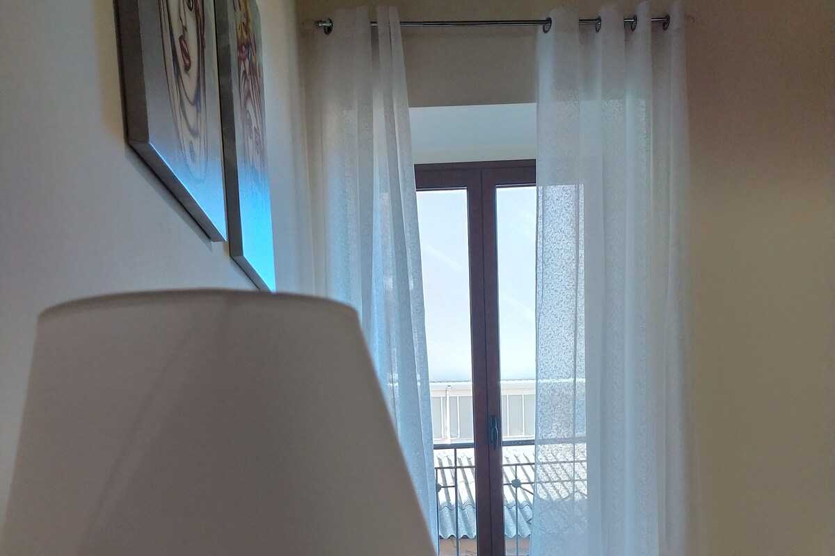 A room with a bed, a lamp on a bedside table, and a view of a balcony through a window with sheer curtains.