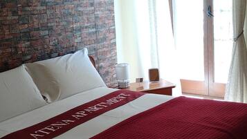 Una camera d'albergo con un letto grande, una testiera con motivo a mattoni, una coperta rossa, un comodino con una lampada e una finestra con tende.