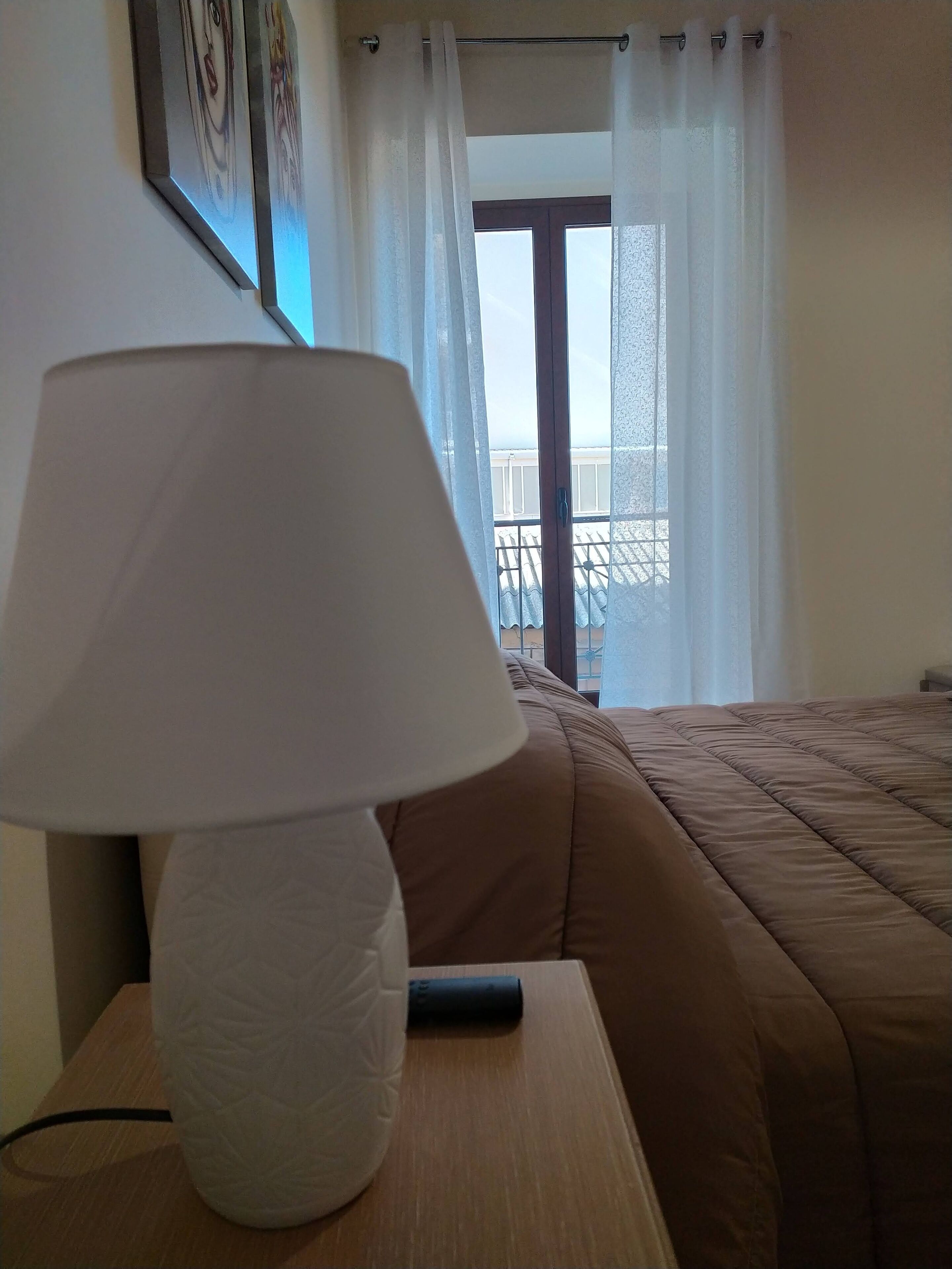 Ett hotellrum med en säng, en sänglampa, ett fönster med gardiner och utsikt från balkongen.