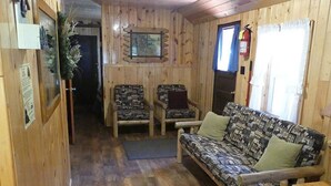 En inredning med träpanel, en soffa, två mönstrade fåtöljer, ett träbord och en dörr som leder till ett annat rum.