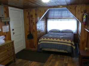 Un dormitori de cabana de fusta amb un llit, una petita còmoda, una finestra i un extintor.