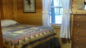 A wooden cabin room with a bed, a dresser, and a window with curtains. - Madison River Cabin Rental #9 (W YELLOWSTONE)