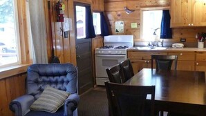 A wooden cabin interior with a dining table, chairs, a fireplace, a stove, and a window with curtains. - Madison River Cabin Rental #9 (W YELLOWSTONE)