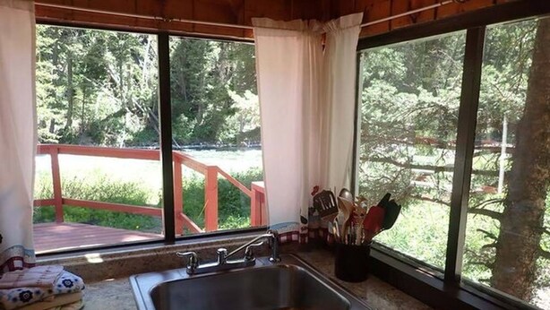 A kitchen with a stainless steel sink, a window with a view of a wooden deck and greenery, and a window blind. - Madison River Cabin Rental #12 (W YELLOWSTONE)
