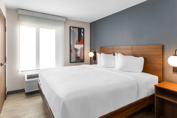 A modern hotel room with a large bed, a bedside table, a wall-mounted lamp, and a window with blinds.