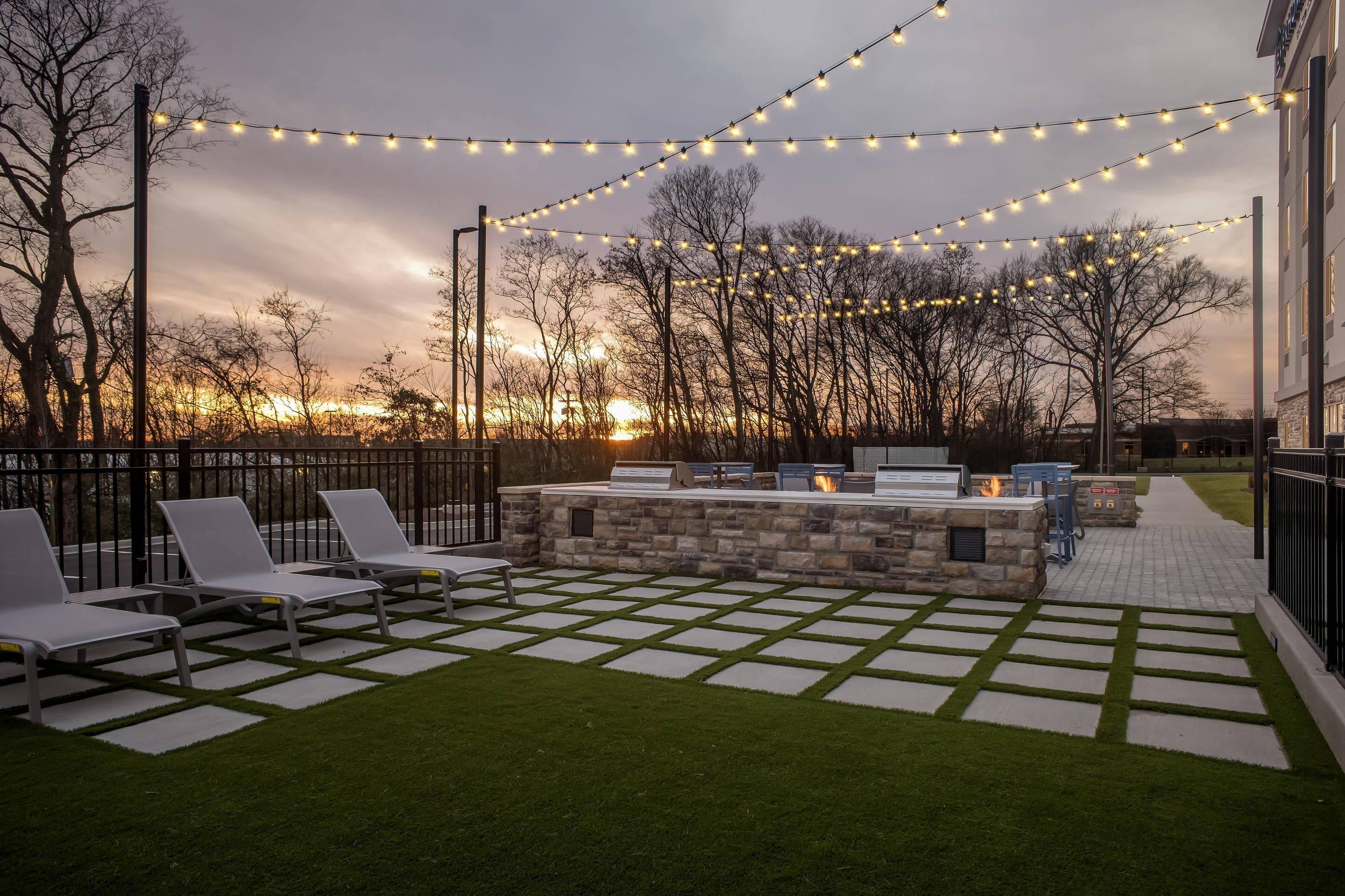 An outdoor seating area with a stone fire pit, lounge chairs, and string lights against a sunset backdrop.
