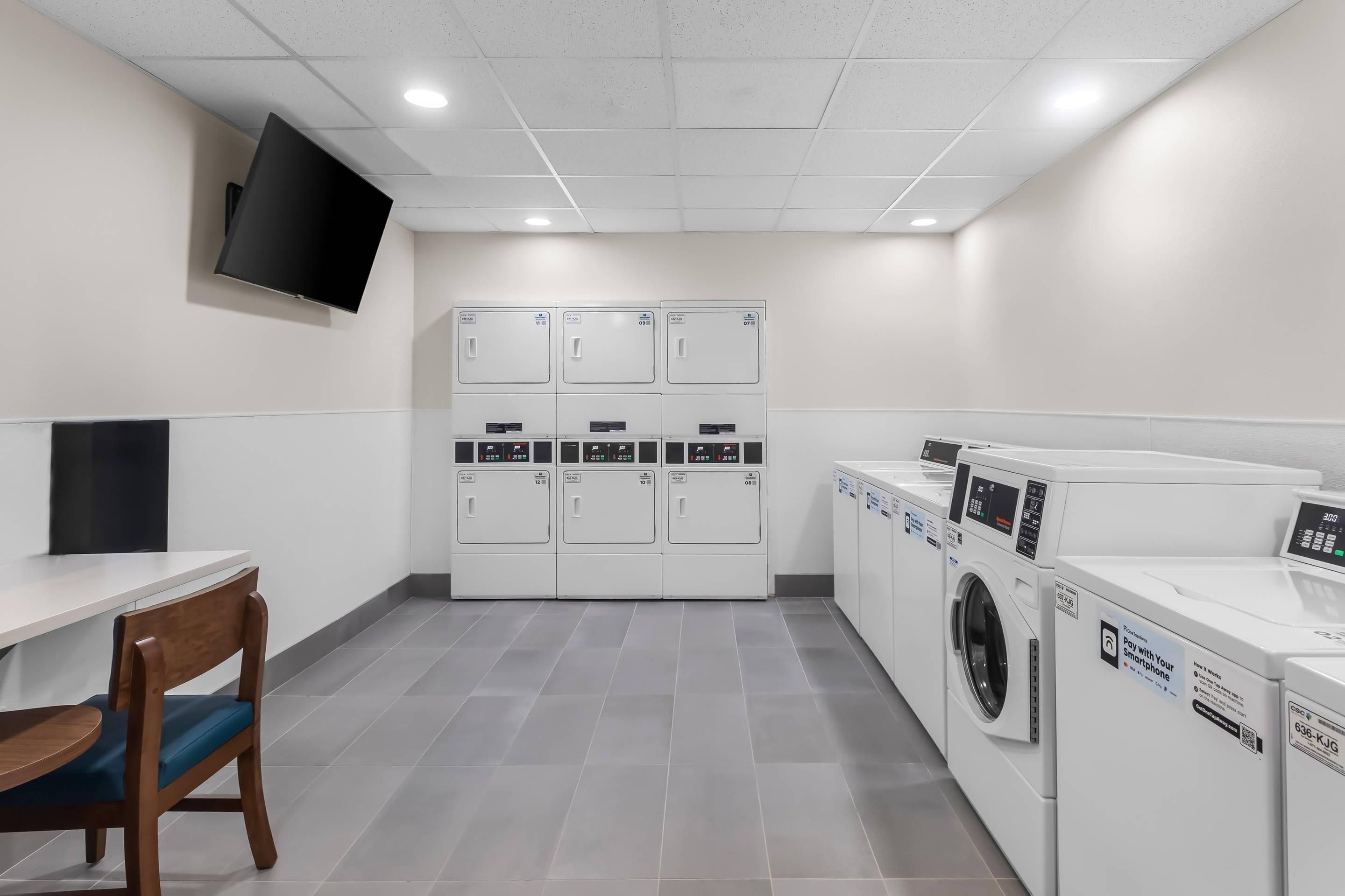 A laundry room with multiple washing machines and dryers, a flat-screen TV mounted on the wall, and a wooden chair with a blue cushion.