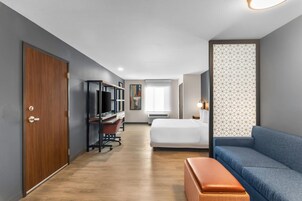 A hotel room with a wooden door, a desk with a computer, a bed, a blue sofa, and a patterned wall.