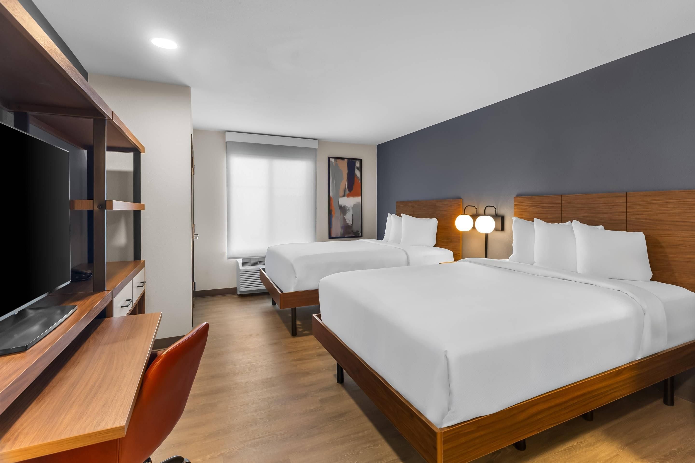 A hotel room with two beds, a desk, a TV, and a large window.