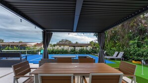 Une terrasse extérieure avec une table et des chaises en bois, un espace salon couvert et une vue sur une piscine et la verdure.