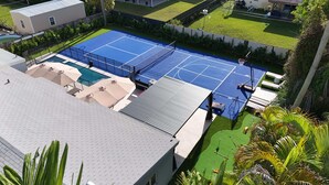 Un court de tennis situé sur le toit d’un bâtiment, avec une piscine, le tout entouré d’une clôture et de verdure.