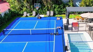 Un court de tennis bleu avec un filet, entouré d’une piscine et d’un espace de détente extérieur.