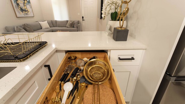 Une cuisine avec un plan de travail blanc, un tiroir contenant des ustensiles de cuisine, un canapé gris et une porte blanche à l’arrière-plan.