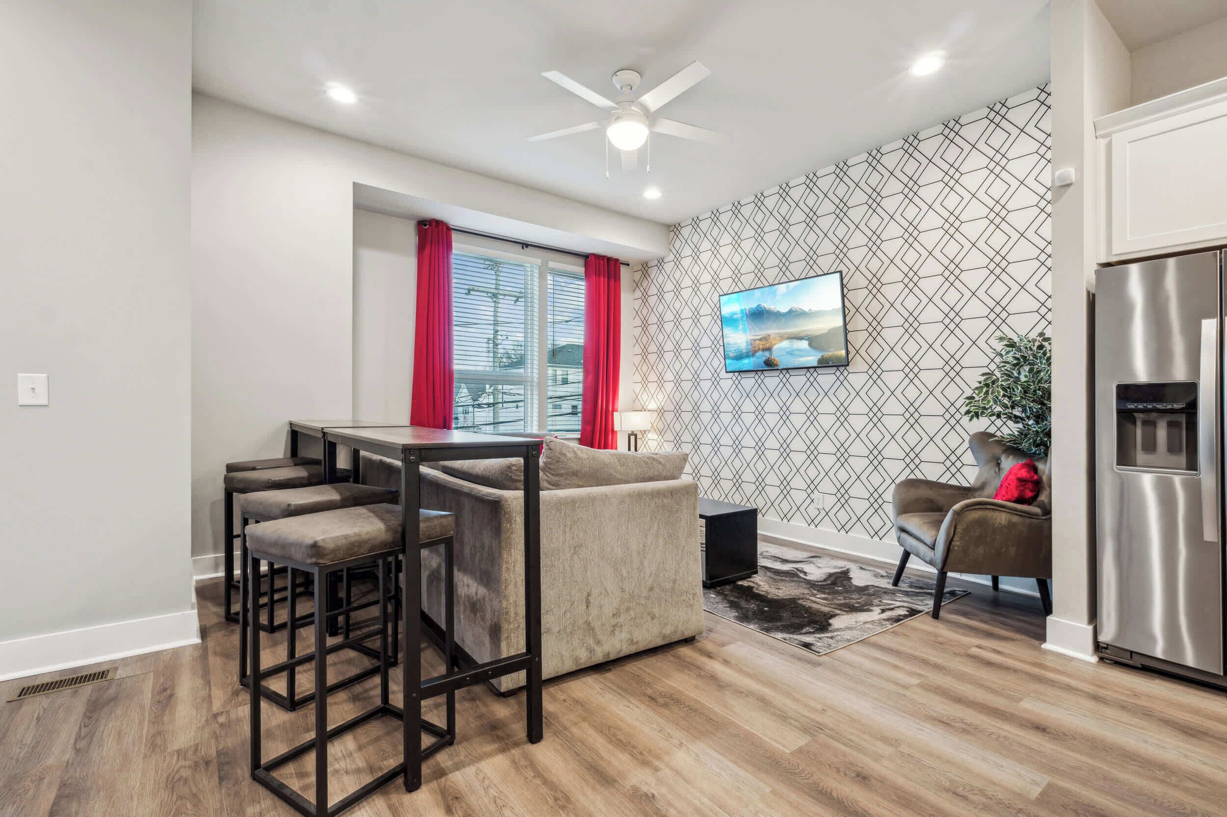 A modern kitchen with a stainless steel refrigerator, a dining area with a bar stool set, a sofa, and a TV mounted on the wall.