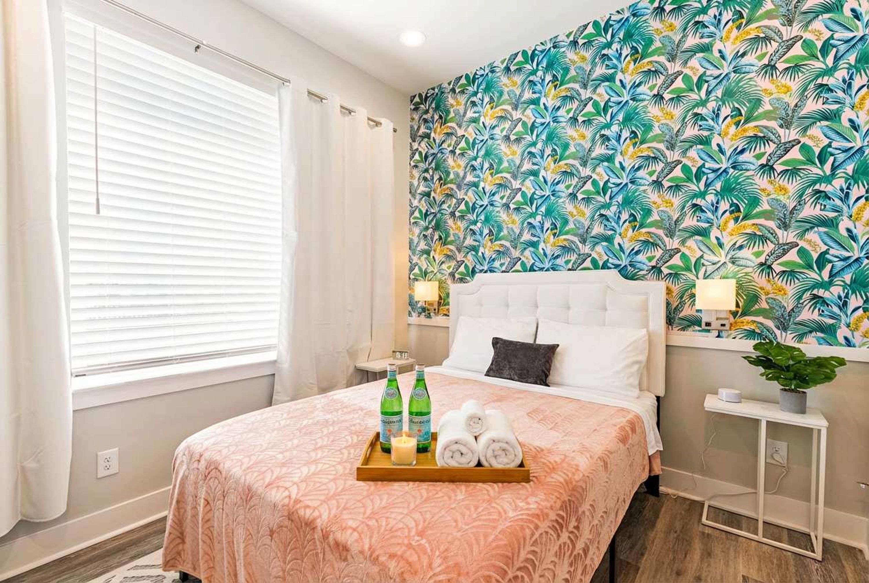 A hotel room with a bed, a tropical wallpaper, a tray with bottles and towels, and a bedside table with a plant.