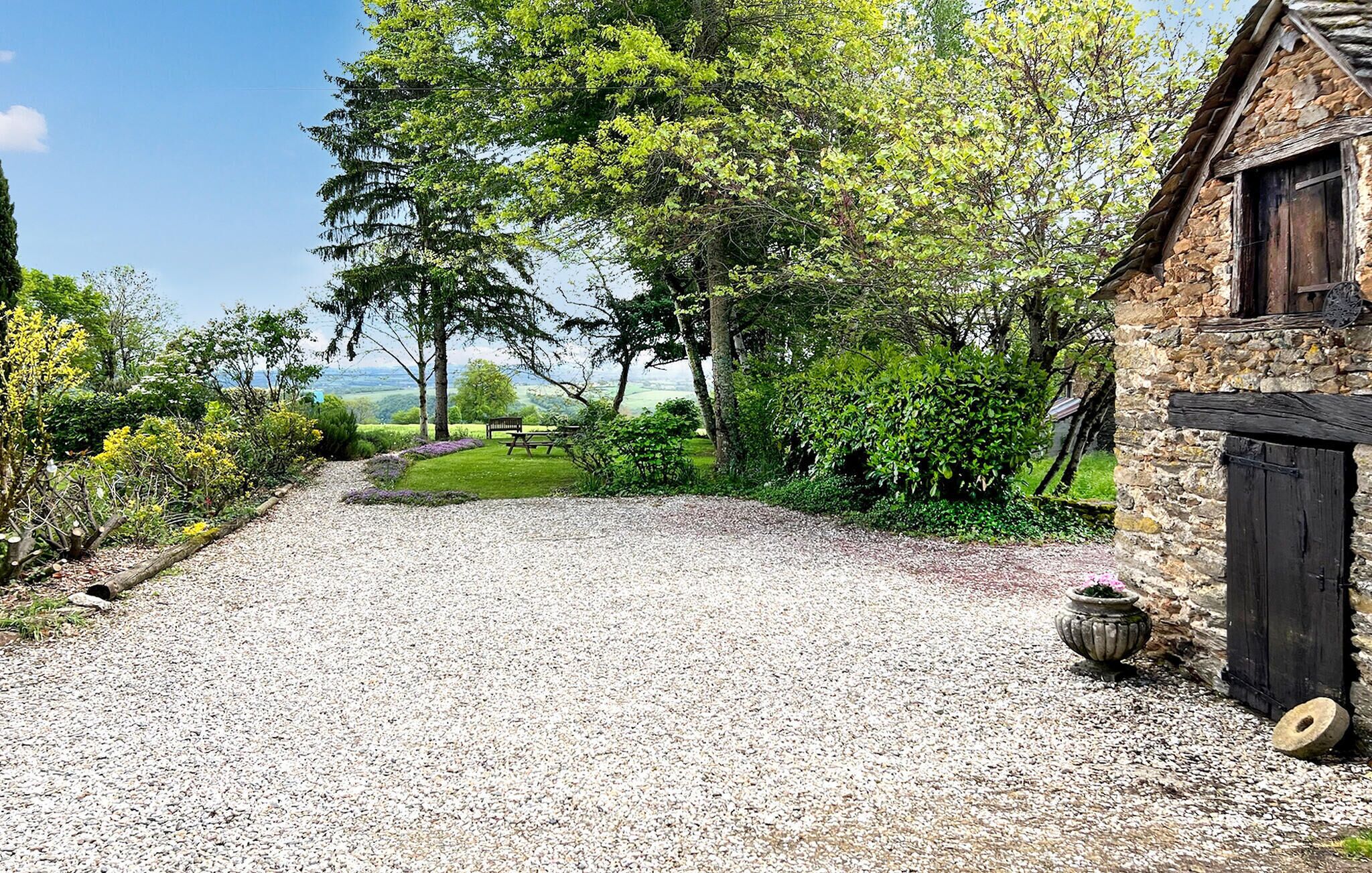 A gravel driveway leading to a stone building with a wooden door, surrounded by lush greenery and a bench.