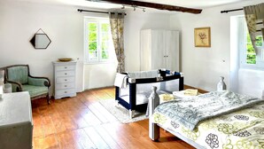 A bedroom with a wooden floor, a bed with a floral bedspread, a dresser, a chair, a crib, and a window with curtains. - 3 bedroom amazing home in Jouqueviel (Jouqueviel)