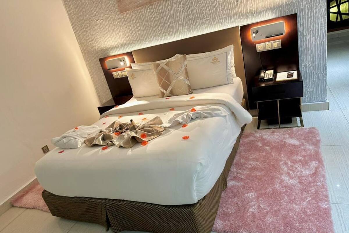 A neatly made bed with a white comforter and pillows, a pink rug, and a bedside table with a phone and a small lamp.