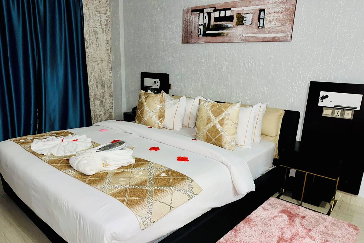 A hotel room with a bed, pillows, a bedside table, a nightstand, a wall clock, and a painting.