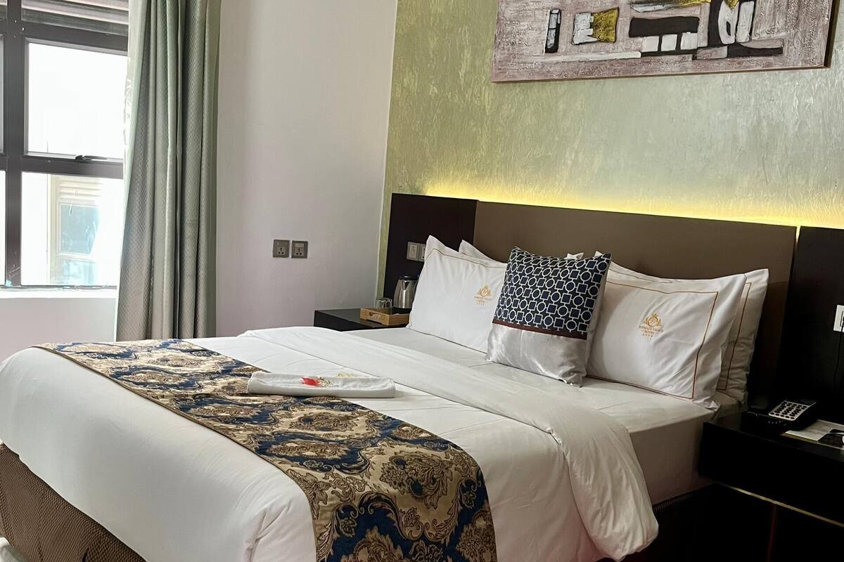 A hotel room with a large bed, a headboard with a light fixture, a nightstand with a phone, and a window with curtains.