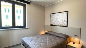 Une chambre à coucher moderne avec un lit, des tables de chevet, un luminaire au plafond, une fenêtre et une œuvre d’art accrochée au mur.