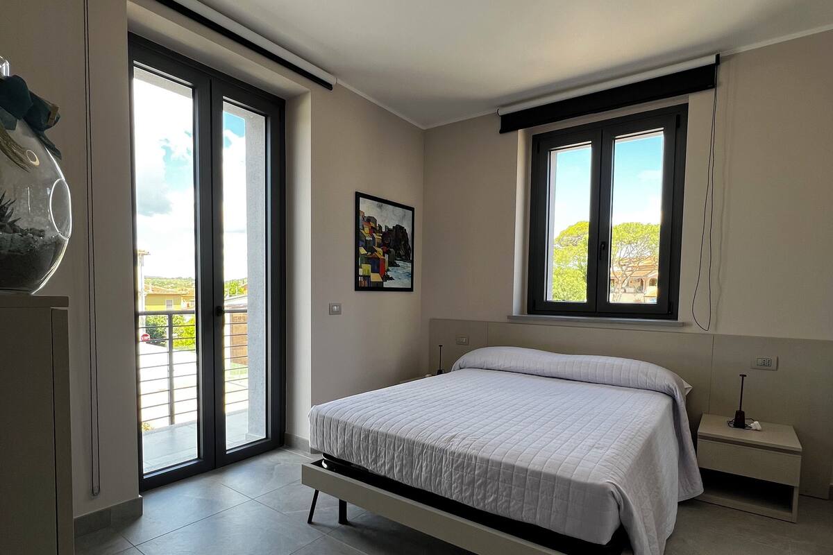 A bedroom with a bed, a nightstand, a wall-mounted lamp, a window with a view, and a ceiling light fixture.