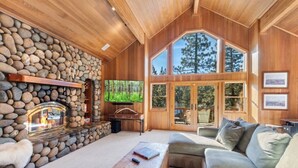 A cozy living room with a stone fireplace, wooden walls, and large windows offering a view of trees. - Northstar Summer Hub | Hot Tub | MTB & Golf Access (Placer County)
