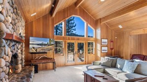 A log cabin interior with a stone fireplace, a large window, and a cozy seating area. - Northstar Summer Hub | Hot Tub | MTB & Golf Access (Placer County)