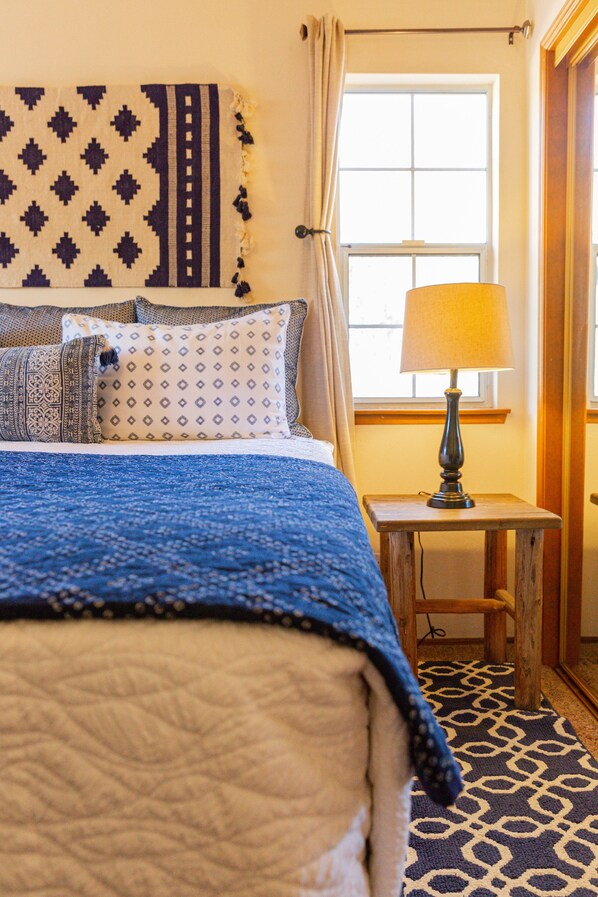 Una cama con un edredón azul, almohadas estampadas y una mesita de noche de madera con una lámpara.