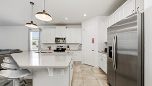A modern kitchen with white cabinets, a stainless steel refrigerator, a central island with bar stools, pendant lighting, and a microwave. - Gated 7BR Resort Villa w/ Private Heated & Lighted Pool | Corner Lot Near Disney (Kissimmee)