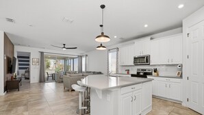 A modern kitchen with white cabinetry, a central island with bar stools, a microwave, and a ceiling fan. - Gated 7BR Resort Villa w/ Private Heated & Lighted Pool | Corner Lot Near Disney (Kissimmee)