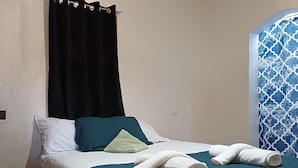 A neatly made bed with a teal bedspread, white pillows, and a green pillow. There are two rolled white towels on the bed. - La Belle Vue Guest House  (Aït Benhaddou)