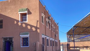 A building with multiple windows and doors, a covered area with a blue roof, and a clear blue sky. - La Belle Vue Guest House  (Aït Benhaddou)