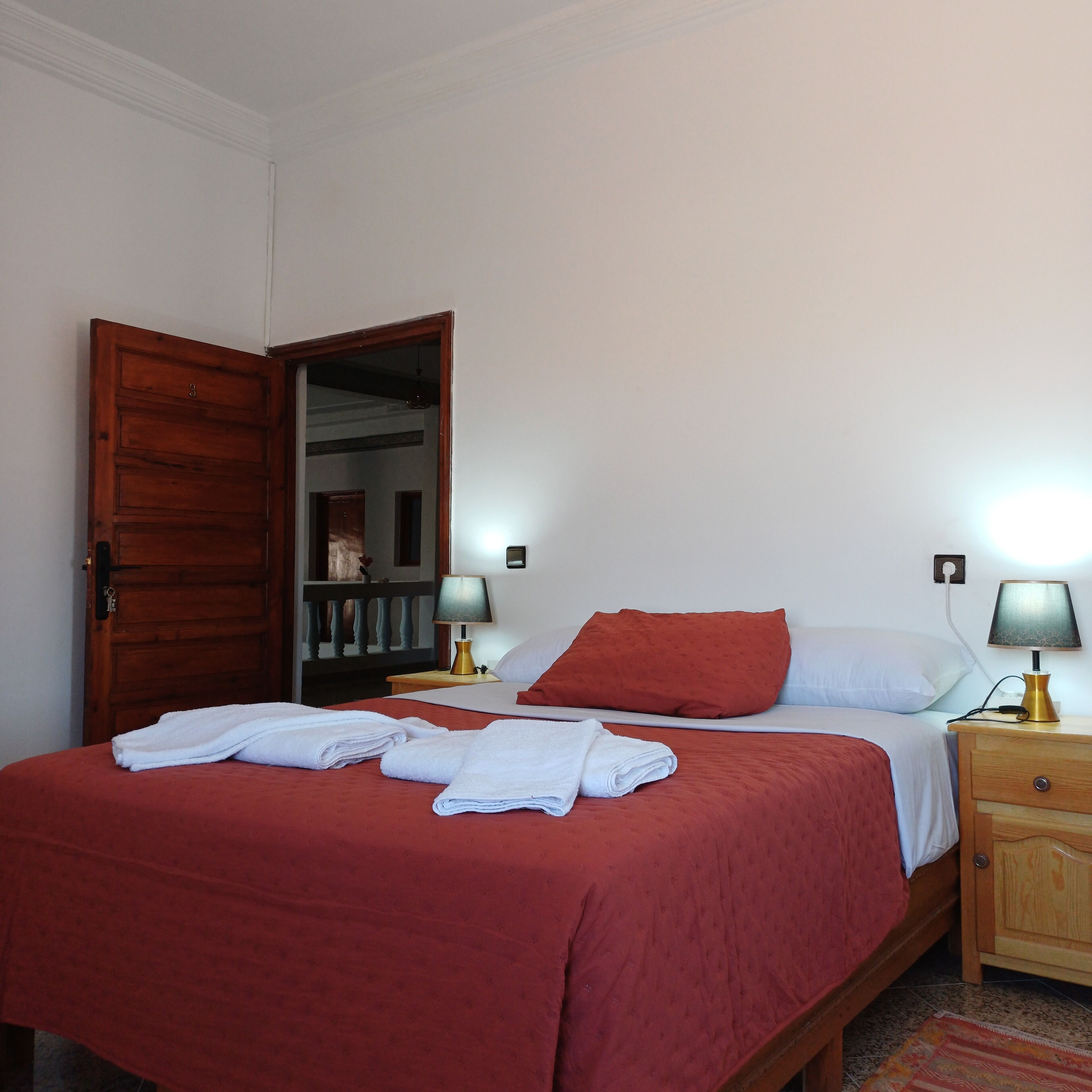 A bedroom with a wooden door, two bedside lamps, a bed with red bedding, and a small wooden nightstand.