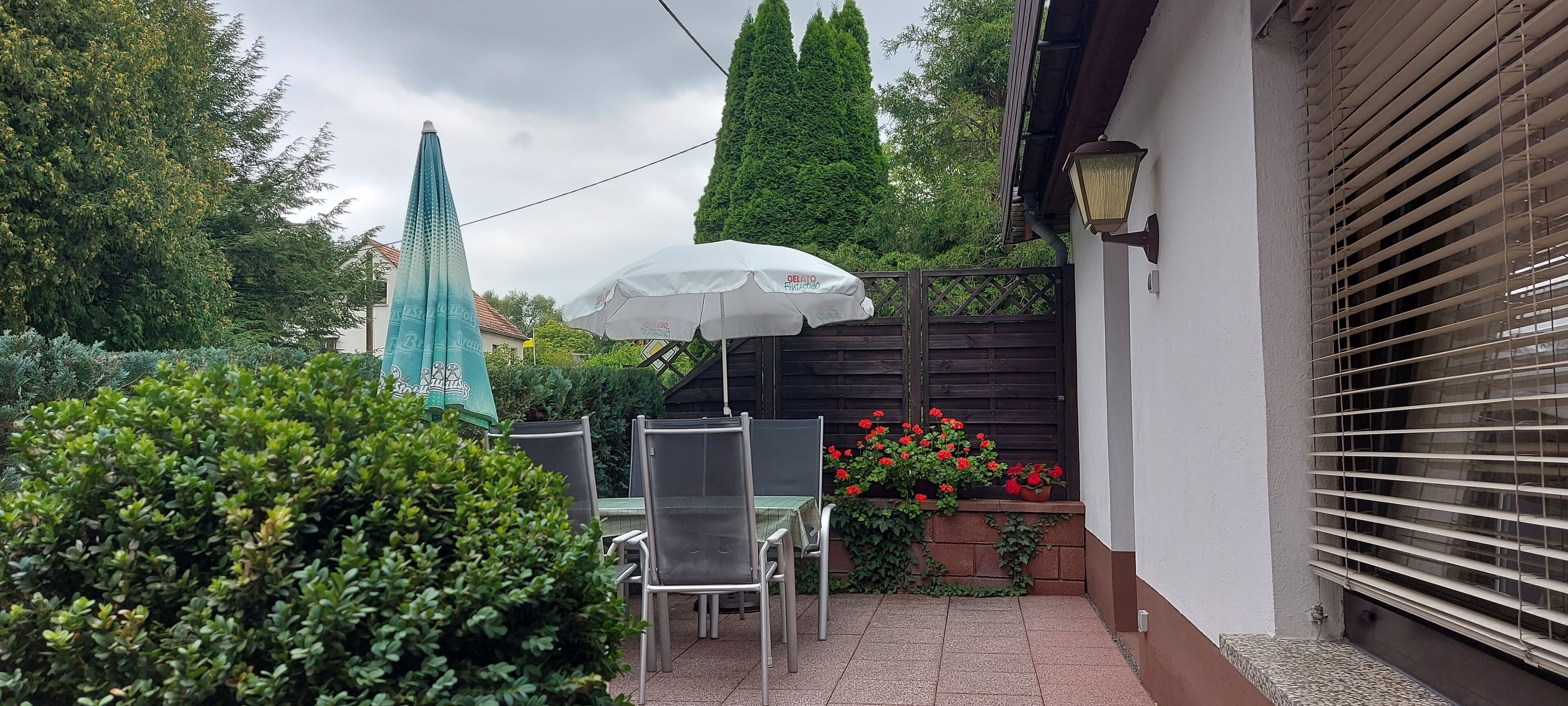 Eine Terrasse mit Tisch und Stühlen, einem weißen Sonnenschirm und einem Ziegelpflanztopf mit roten Blüten.