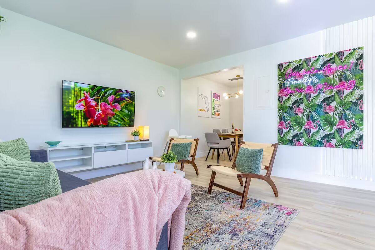 A modern living room with a flat-screen TV, a sofa, a dining area, and a large window with a tropical print.