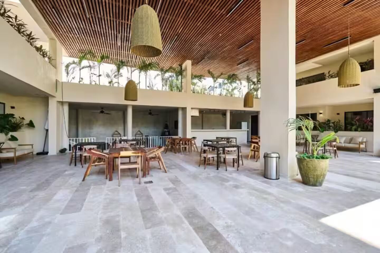 Un espacio interior amplio con varias mesas y sillas para comer, grandes lámparas colgantes y un techo alto con listones de madera.