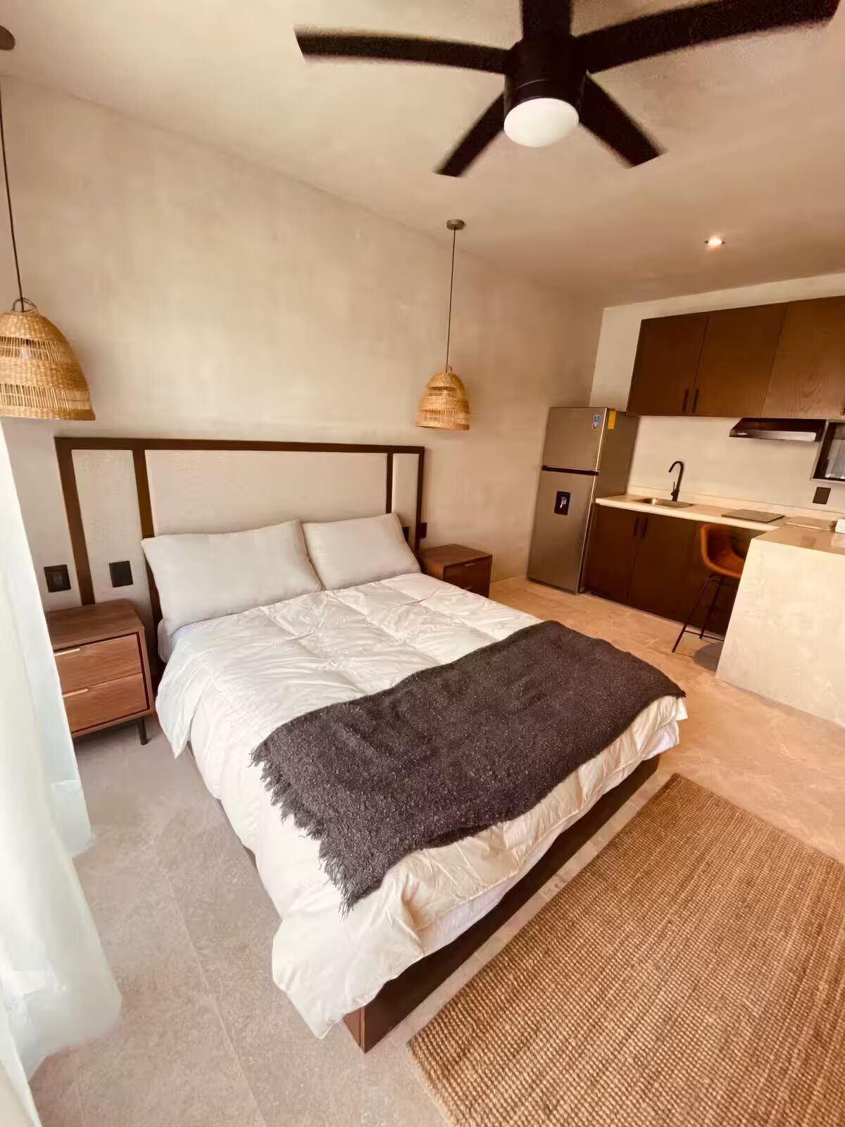 A bedroom with a bed, bedside tables, a ceiling fan, and a kitchenette in the background.