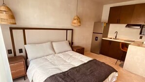 Un dormitorio con cama, mesitas de noche, ventilador de techo y una kitchenette al fondo.