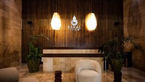 Zona de recepción con pared de paneles de madera, dos lámparas colgantes, un mostrador de recepción, un sillón blanco y dos plantas grandes en macetas.