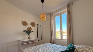 A bedroom with a bed, a bedside table, a hanging light fixture, a window with curtains, and a wall-mounted mirror. - Joli t2 (Marius) (Aubagne)