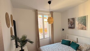 A bedroom with a bed, a small table with a plant, a hanging light fixture, and a window with curtains. - Joli t2 (Marius) (Aubagne)