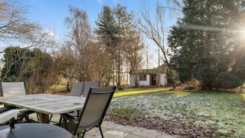 Um pátio externo com mesa e cadeiras, um pequeno galpão e um jardim com árvores e gramado.