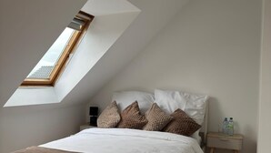 Quarto com teto inclinado, claraboia, cama com manta marrom, roupa de cama branca e mesa de cabeceira com garrafas de água.