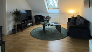 Um loft espaçoso com pé-direito alto, vigas de madeira, um amplo claraboia, televisão de tela plana, sofá, mesa de centro e um vaso com planta.