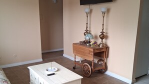Un salon avec un meuble blanc, un canapé à motifs floraux, un chariot en bois sur lequel sont posées des lampes, et un téléviseur à écran plat fixé au mur.