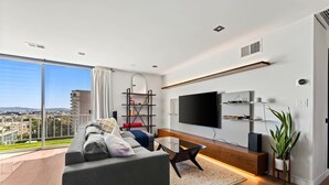 A modern living room with a large sofa, a coffee table, a TV mounted on the wall, and a balcony with a city view. - Modern Cozy 1-bedroom condo in Beverly Hills (Los Angeles)
