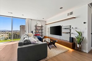 Ein modernes Wohnzimmer mit einer großen Couch, einem Couchtisch, einem an der Wand befestigten Fernseher und einem Balkon mit Blick auf die Stadt.